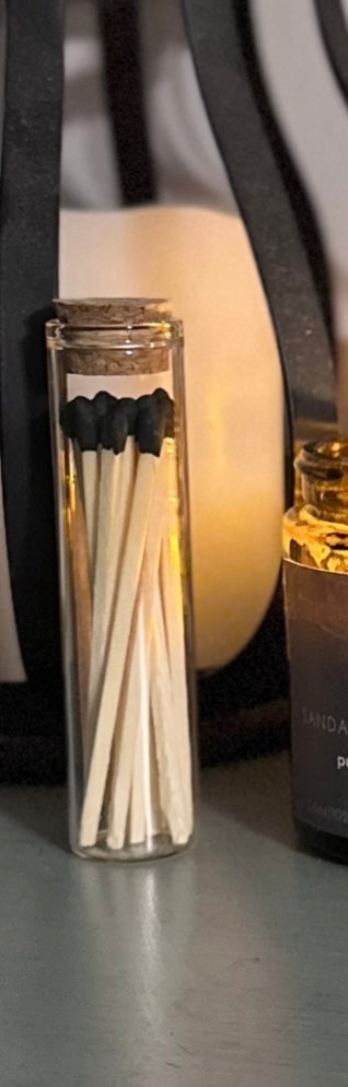 Glass container of matches with cork lid next to a candle lit on a shelf